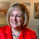 Cheryl Fishbein with blond hair, wearing a bright red jacket and a beaded necklace, is smiling. She is sitting in a warmly lit room with wall art in the background. The 2023 Manhattan Jewish Hall of Fame Inductee exudes warmth and charm.