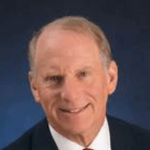 Dr. Richard Haass with short, light brown hair, wearing a dark suit jacket, white dress shirt, and a dark tie. The 2023 Manhattan Jewish Hall of Fame Inductee is smiling and posed against a solid, dark blue background.