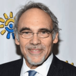 Dr. Irwin Redlener with gray hair and glasses is smiling, wearing a dark suit, a white shirt, and a blue tie. As a 2023 Manhattan Jewish Hall of Fame Inductee, he stands proudly in front of a backdrop with colorful sunburst designs.