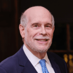 Harold Holzer with a bald head and white beard smiles at the camera. He is wearing a navy blue suit, white shirt, and light blue tie. The background is dark and out of focus, highlighting his distinguished presence as a 2023 Manhattan Jewish Hall of Fame Inductee.