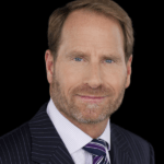 Portrait of Kent Swig with short light brown hair and a beard, wearing a dark pinstripe suit, a light pink checkered dress shirt, and a purple striped tie. The background is black, and he is smiling slightly while looking directly at the camera—a 2023 Manhattan Jewish Hall of Fame inductee.
