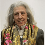 Patti Askwith Kenner with long, wavy, gray hair smiles softly. She is wearing pearl earrings and a colorful scarf with gold, pink, and black patterns draped over a dark jacket and white blouse. This 2023 Manhattan Jewish Hall of Fame Inductee stands against a plain white background.