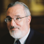 A middle-aged man with short, gray hair and a beard is wearing glasses, a suit, and a tie. He is looking slightly to the side, with a neutral expression on his face. The background is softly blurred, drawing focus to his face—an image befitting a 2023 Manhattan Jewish Hall of Fame Inductee.