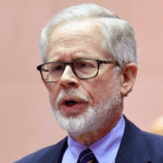 A man with short white hair and a white beard is wearing glasses, a dark suit, a light blue dress shirt, and a striped tie. He appears to be speaking or addressing an audience as the 2023 Manhattan Jewish Hall of Fame Inductee. The background is a soft, warm color.